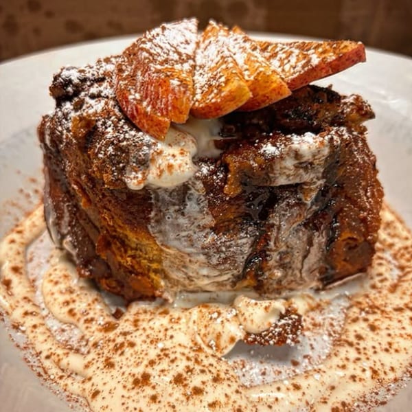 Bread pudding with powdered sugar, cream, cinnamon, and orange slices on a white plate.