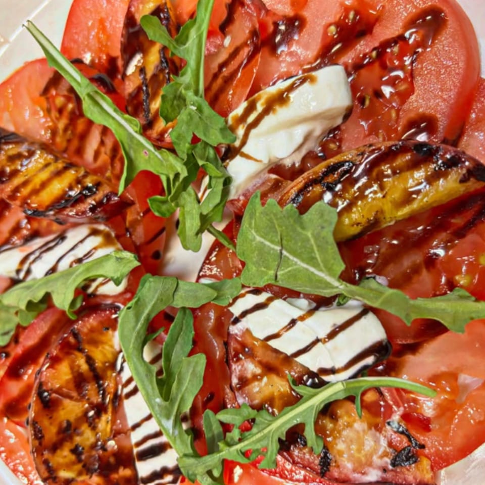 Close-up of a Caprese salad with sliced tomatoes, grilled peaches, mozzarella, arugula, and balsamic glaze.