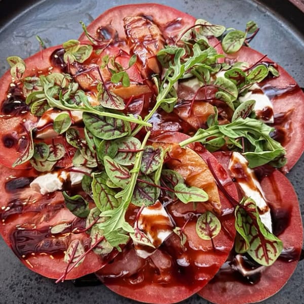 Heirloom tomato carpaccio with fresh mozzarella, microgreens, and balsamic glaze on a dark plate.