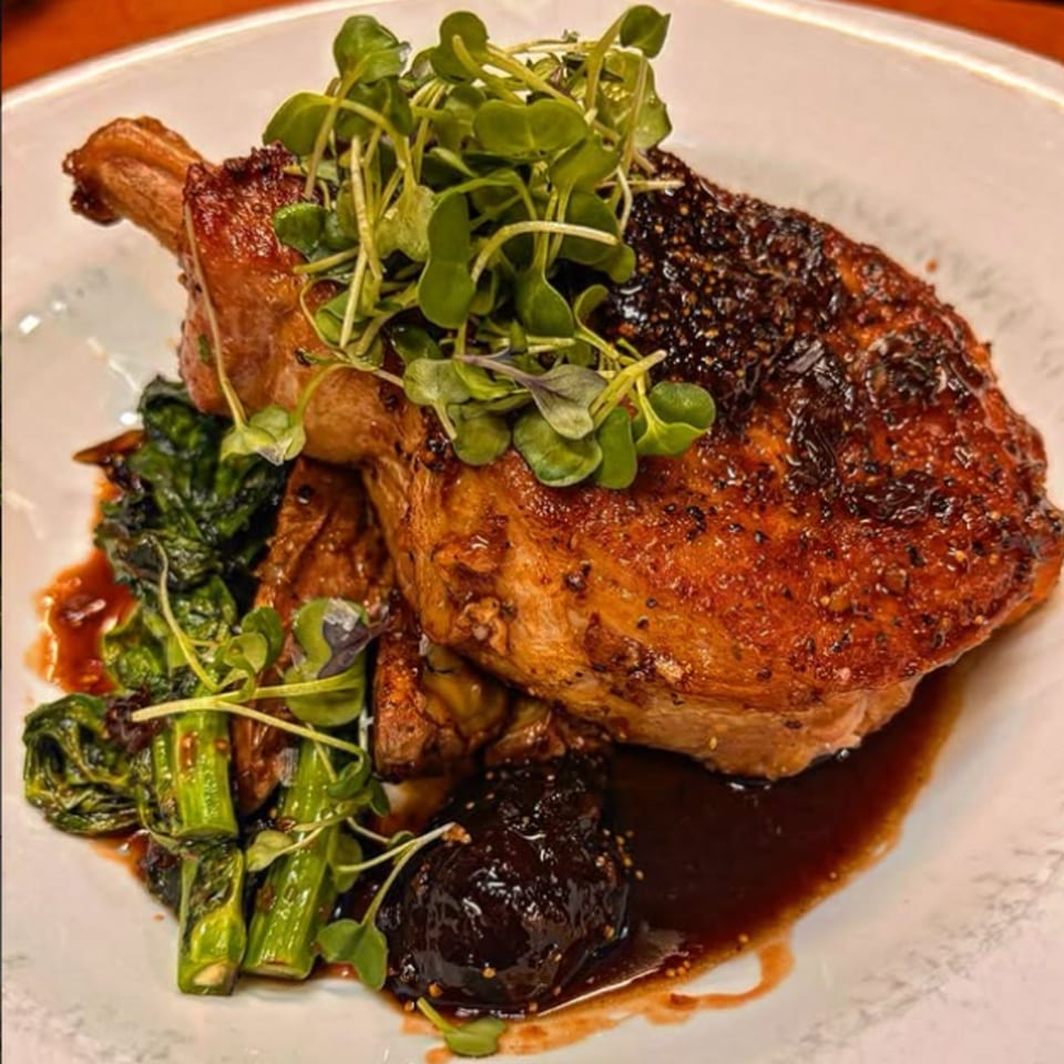 Crispy duck leg confit with dark sauce, green vegetables, and microgreens on a white plate.